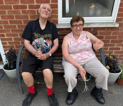 Two smiling adults sit on a bench outside a brick building, surrounded by potted plants.